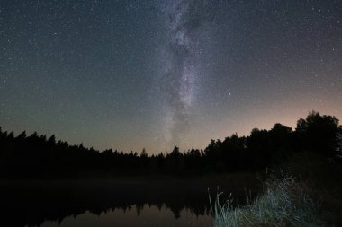 Samanyolu, ormanın karanlık bir siluetiyle çevrili gölün ayna yüzeyine yansımaktadır. Estonya 'da sakin bir gece.