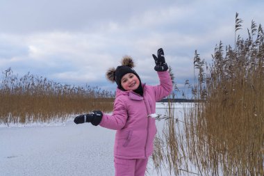 A little girl, a child of 7 years old, stands on the shore of a frozen lake, Harku, on a cloudy winter day. clipart