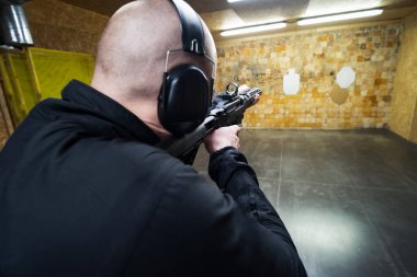 Kel adam atış poligonunda tüfeğini ateşliyor, arkadan fotoğraf çekiyor..