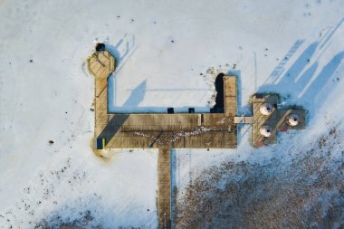 Drone view of a frozen pier with gazebos, surrounded by snow: long shadows, deserted winter landscape, cold silence.