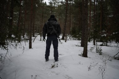 Siyah üniformalı bir asker, sırtında makineli tüfekle, alacakaranlıkta bir çam ormanında, arkadan bakıyor..