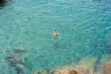 Bir kız mavi şeffaf suyla denizde şişme bir yüzükte yüzüyor..