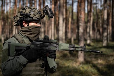 Elinde tüfeği olan miğferli bir asker, çam ormanında nöbet tutuyor, etrafa bakıyor..