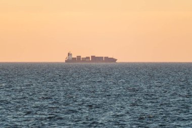 Konteynırları olan büyük bir kargo gemisi mavi denizde gün batımında sıcak turuncu bir gökyüzünün altında yelken açar..