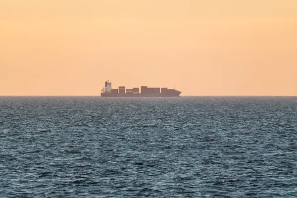 Konteynırları olan büyük bir kargo gemisi mavi denizde gün batımında sıcak turuncu bir gökyüzünün altında yelken açar..