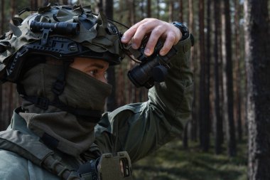 Kamuflajlı asker ve miğfer, ormanda devriye gezerken gece görüş cihazını ayarlar..