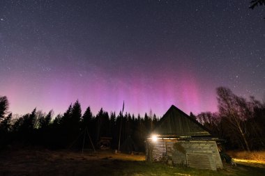 Estonya 'daki köy kulübesi göz kamaştırıcı pembe ve mor aurora borealis ve yıldızlı gece gökyüzünün altında sıcacık bir şekilde parlıyor..