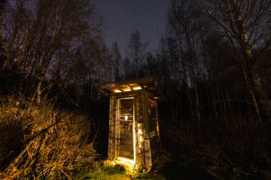 Eski ahşap bir ek bina gizemli bir şekilde uzak yıldızların altındaki karanlık gece ormanında sıcak ışıkla parlıyor..