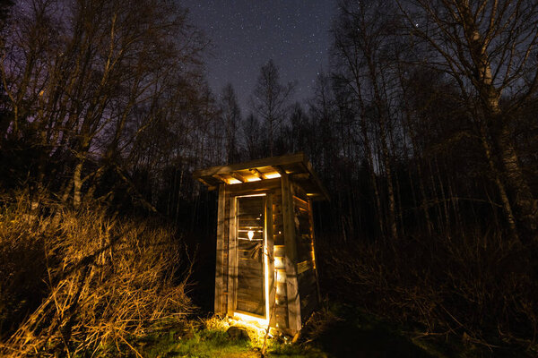 An old wooden outhouse mysteriously glows with warm light in the dark night forest under distant stars.