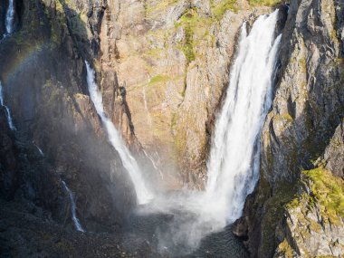 Güçlü Voringfossen şelalesi, Norveç: Güneş ışığı altındaki kayaların arasına düşen etkileyici bir su manzarası, gökkuşağı parıltısıyla.