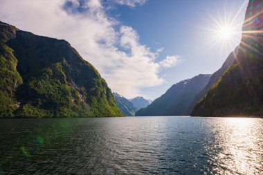 Norveç 'in nefes kesici doğası: görkemli yeşil dağlar fiyortun sakin sularını parlak güneş ve mavi gökyüzü altında kucaklıyor.