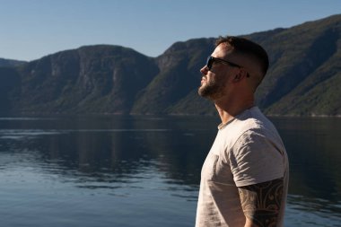 Kolunda dövmesi olan güneş gözlüklü bir adam sıcak güneşin ve Norveç fiyortunun huzurlu manzarasının tadını çıkarır..