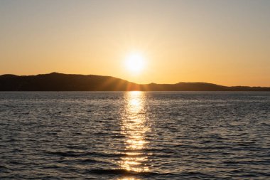 Norveç 'in sakin suları üzerinde altın bir günbatımı, yüzeye yansıyor ve uzak sahillerin siluetlerini aydınlatıyor..