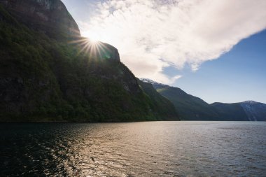 Norveç fiyordunun üzerindeki sihirli ışık: Güneş yüksek karanlık bir dağın arkasından doğar, su yüzeyindeki parlak parlaklıkları resmeder ve bulutları aydınlatır.