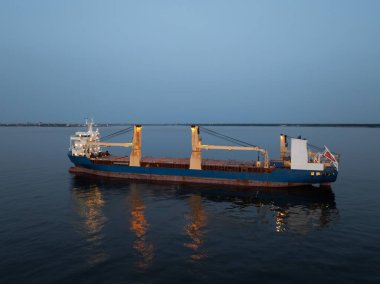 Büyük çapalı bir kargo gemisi. Işıkları alacakaranlığı delip geçiyor. Sakin bir akşam gökyüzünün altındaki sakin sulara yansıyor..