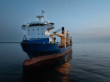 Kargo gemisi alacakaranlıkta, Estonya 'da Baltık Denizi' nde. Hilal şeklinde bir ay ve sakin bir gökyüzünün altında ışıkları açık olan bir gemi..