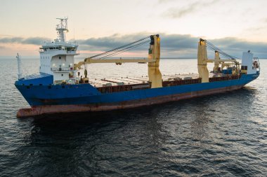 İnsansız hava aracı görüntüsü: Baltık Denizi 'nin sakin sularında gün batımında resim gibi bir gökyüzünün altında sarı turnalı mavi bir kargo gemisi..