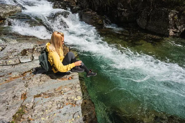Sırt çantalı bir kadın turist Norveç 'teki çalkantılı bir dağ nehrinin yanındaki bir kayanın üzerinde dinleniyor. Yaz yürüyüşü, macera ve vahşi doğayla bağlantı kavramı.