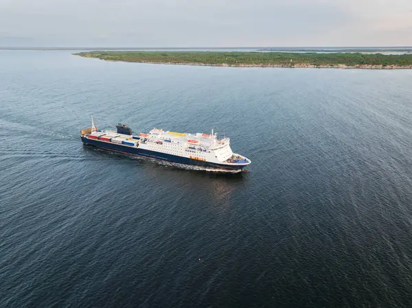 Konteynır ve araçlar taşıyan büyük bir kargo yolcu feribotu, arka planda resim gibi yeşil bir adayla sakin bir denizde yelken açıyor. Bir İHA 'dan havadan çekim.