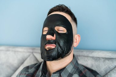 Kapalı gözlü bir adam yüzünde siyah bir maskeyle rahatlar. Erkeklerin cilt bakımı ve spa tedavisi kavramı.