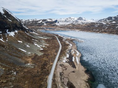 Destansı bir yolculuk: İskandinavya 'daki sert bir dağ geçidinde kısmen donmuş bir göl boyunca dönen manzaralı bir yolun insansız hava aracı görüntüsü.