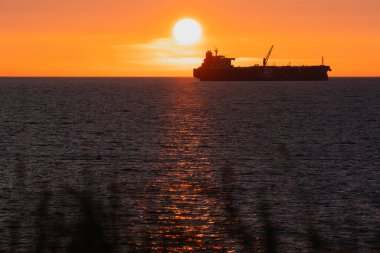 Dev bir geminin silueti, Baltık Denizi 'nin sakin sularında kızgın bir günbatımında ufuk çizgisini ve suyun üzerindeki güneş yolunu yavaşça geçer..