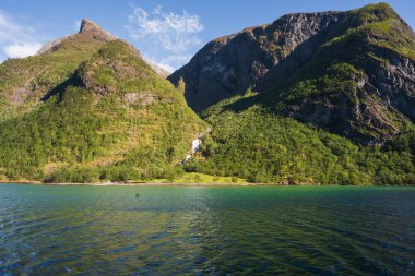 Işıl ışıl bir yaz gününde, ikonik Norveç manzarası derin bir fiyort ve şelale ile yemyeşil dağlar içerir..
