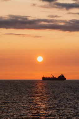 Ufukta görünen bir kargo gemisi silueti altın bir günbatımında, parlak bir güneş yolu ile denize yansıyor..