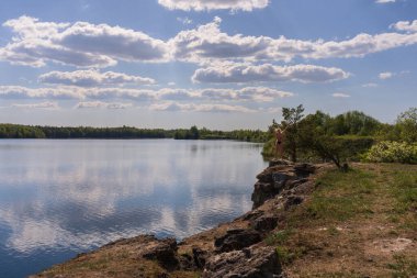 Güzel bir yaz manzarası, bulutların yansımasıyla sakin ve sakin bir göle bakan kayalık bir uçurumda duran bir insanı gösteriyor.. 