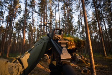 Birinci şahıs bakış açısı: taktik teçhizatlı bir asker, orman üzerinde uçan bir drona kırmızı nokta görüşlü bir saldırı tüfeği doğrultuyor..