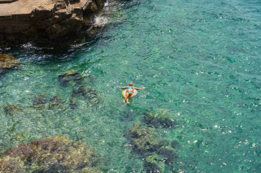 Yazın kayalık sahil kenarında kristal berrak turkuaz denizde şişme bir yüzüğün üzerinde dinlenen bir kadının üst görüntüsü..