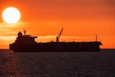 Büyük bir kargo gemisinin silueti, okyanusta yüzen bir petrol tankeri dramatik turuncu gün batımı, deniz ufkunda güneş..