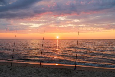 Üç olta kamışı kumlu bir sahilde duruyor, sakin okyanusun üzerinde nefes kesici ve renkli bir gün doğumuna karşı..