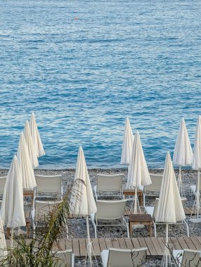 Nice, French Riviera 'nın ikonik çakıl taşı sahilinde sakin bir sabah sahnesi. Kapalı beyaz şemsiyeleri ve boş şezlongları var..