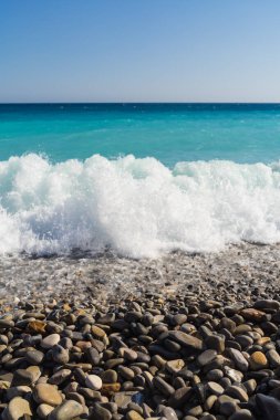 Güzel bir turkuaz okyanus dalgası beyaz sörfle güneşli bir yaz gününde taşlı bir çakıl taşı plajına çarpar..