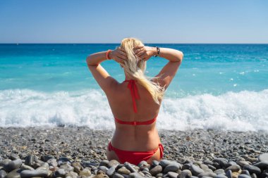 Kırmızı bikinili bir kadının, sarı saçlarını tutarken, turkuaz deniz kıyısındaki çakıl taşı plajında dinlenirken görüntüsü..
