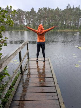 Parlak turuncu yağmurluk giymiş bir kadın ıslak bir ahşap iskelede kollarını açmış yağmuru kucaklıyor..