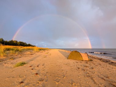 Gün batımında Estonya 'da terk edilmiş bir Baltık Denizi sahilinde, tek kişilik bir kamp alanının üzerinde nefes kesici çift gökkuşağı kemerleri..
