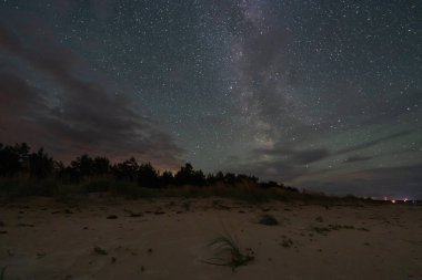 Çarpıcı Samanyolu Galaksisi, karanlık Estonya sahillerinin üzerinde, siluetli ağaçlar ve uzak ışıklarla gece gökyüzünde uzanıyor..
