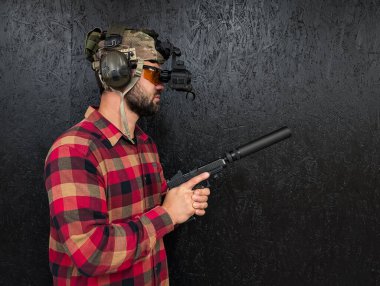 Ekose gömlekli, gece görüşlü ve kulak koruyuculu taktik kasklı bir adam susturuculu bir tabanca tutuyor..