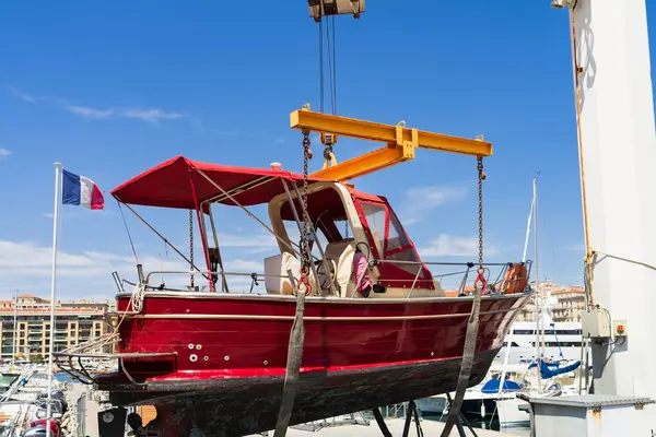 Kırmızı bir motorlu tekne, güneşli bir Fransız limanında bakım ve onarım için büyük sarı bir vinç tarafından kaldırılır..