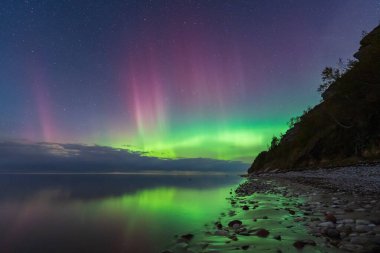 Magical purple and green Aurora Borealis, the Northern Lights, reflecting in the serene sea on a rocky Estonian coast. 