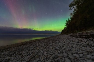 Breathtaking Northern Lights, the Aurora Borealis, shimmer with green and purple hues over a tranquil pebble beach in Paldiski, Estonia.
