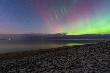 A magical nightscape featuring the vibrant green and purple Aurora Borealis, or Northern Lights, over a tranquil pebble beach in Paldiski, Estonia.