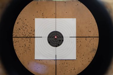 A first-person view through a rifle scope with a red dot sight aimed at a paper bullseye target outdoors.