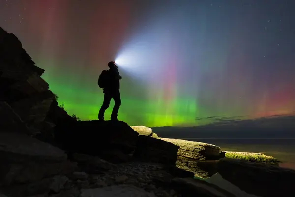 Elinde fener olan maceraperest yürüyüşçü, sihirli Aurora Borealis 'in Estonya' nın kayalık Baltık sahillerinde gece gökyüzünü aydınlatmasını izliyor..