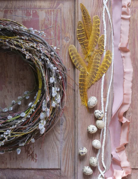 A fragment of an Easter wreath made of tree branches and a pussy willow on a wooden background with decorative elements for Easter: quail eggs, feathers, silk ribbons, decorative braid of sequins that lie nearby
