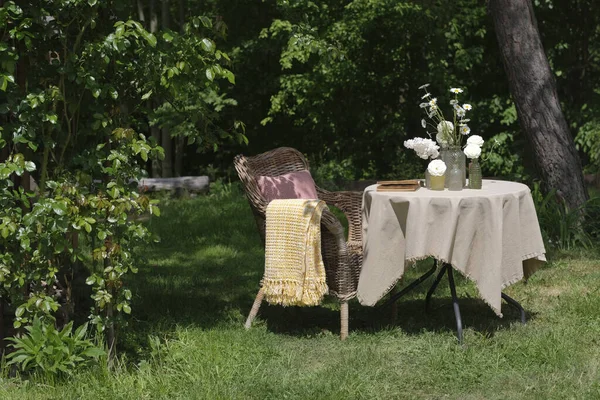 stock image A wicker chair and a table with flowers in the garden on a sunny summer day. A place to relax and read in the fresh air