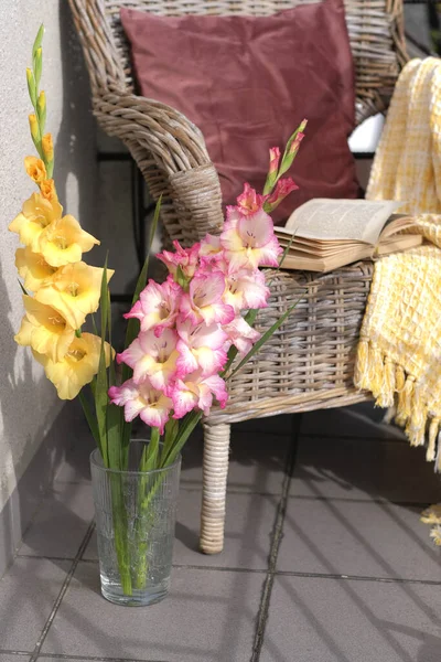 Balkondaki cam vazoda bir buket gladioli, hasır şezlongun arka planında bir yastık ve ekoseli bir sandalye.