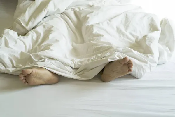 Men\'s feet under a white blanket, in bed, in a comfortable apartment, in natural light, with copy space.
